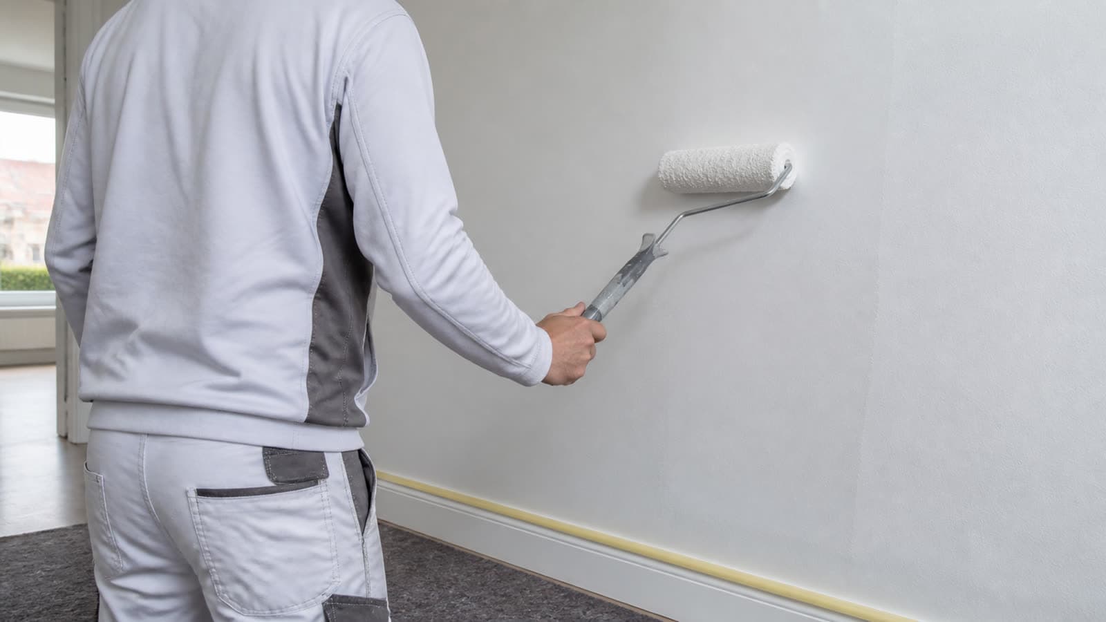 Schilder rolt een lichte muur in een interieur met afplaktape langs de plinten